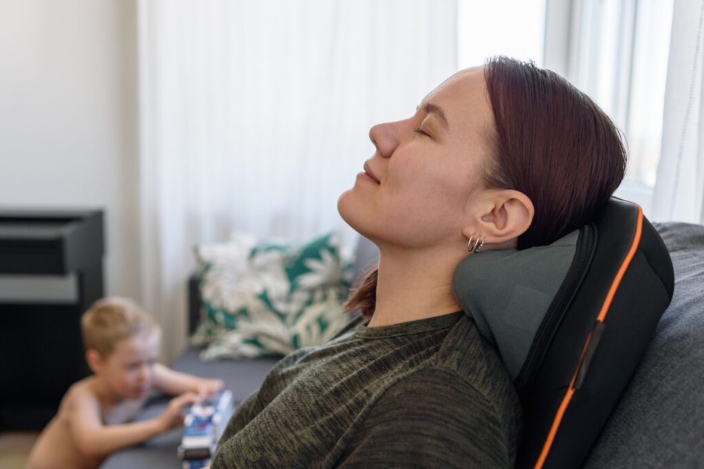 a woman relaxing on a massage chair at home and a child playing beside her